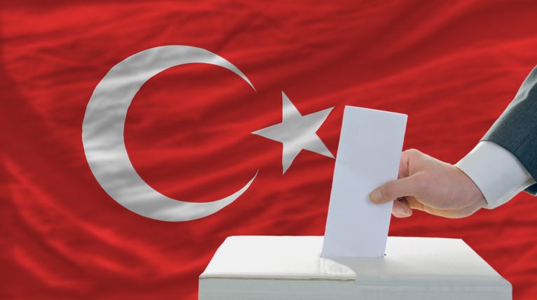 Stimmzettel wird in Wahlurne geworfen, die vor der türkischen Flagge steht