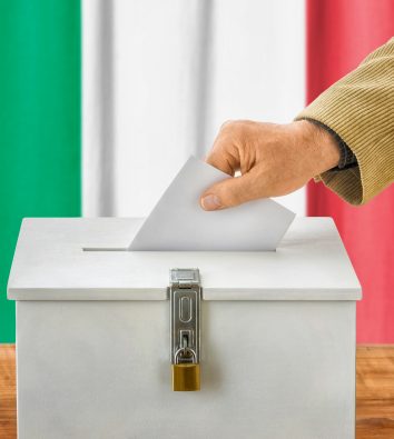 Ein Wahlzettel fällt in die Wahlurne, im Hintergrund die italienische Flagge