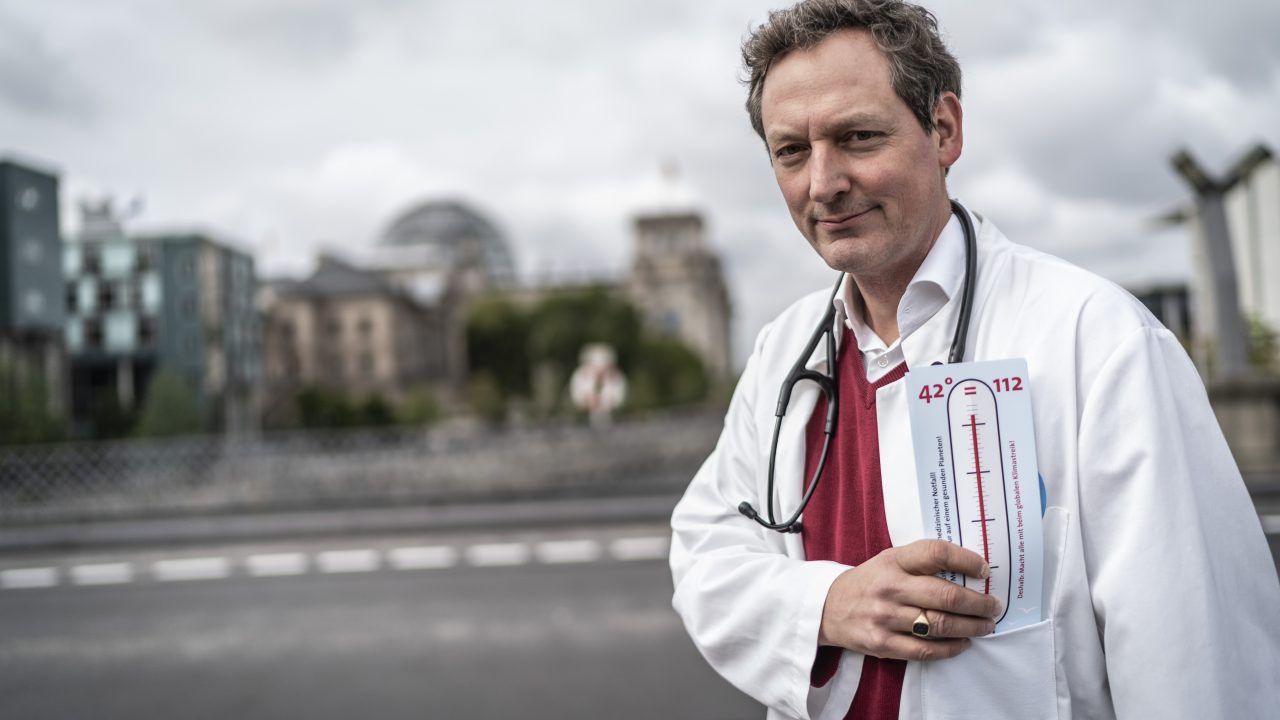Dr. Eckhart von Hirschhausen steht in einem weißen Kittel, mit einem Stethoskop um den Hals und einem Zettel mit einem Fieberthermometer in der Hand vor dem Deutsche Bundestagsgebäude in Berlin.